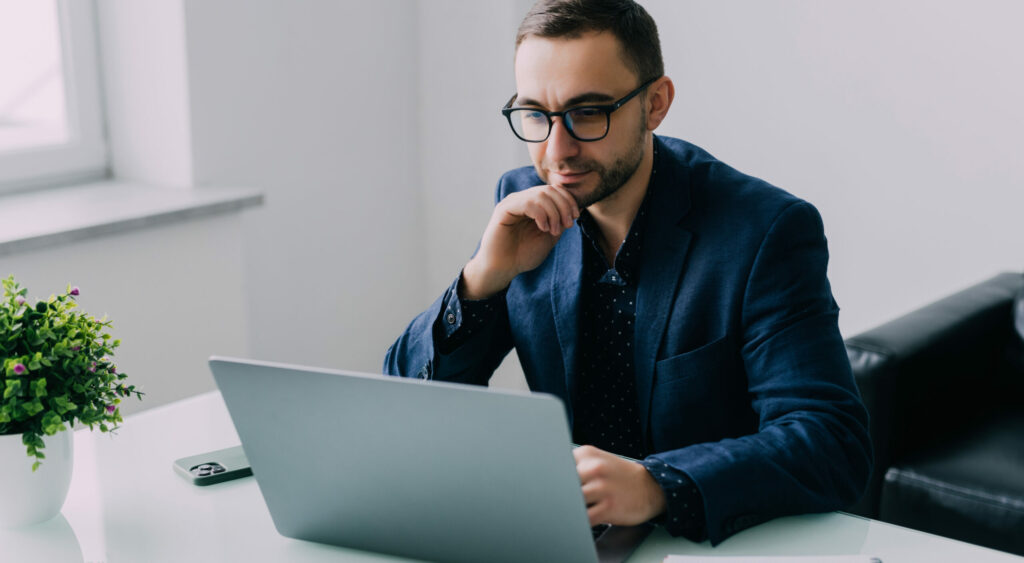 Handsome business executive sitting at his desk in a modern office with windows looking on the screen of his laptop computer