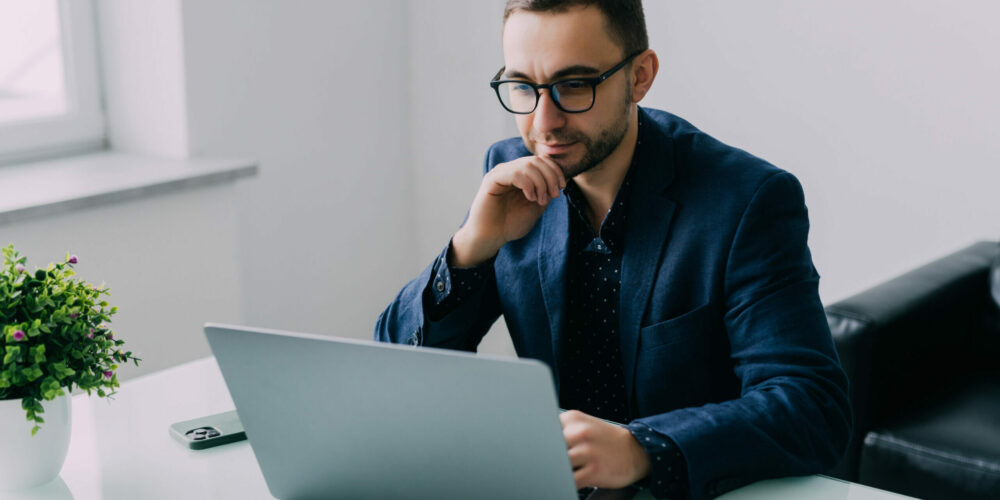 Handsome business executive sitting at his desk in a modern office with windows looking on the screen of his laptop computer